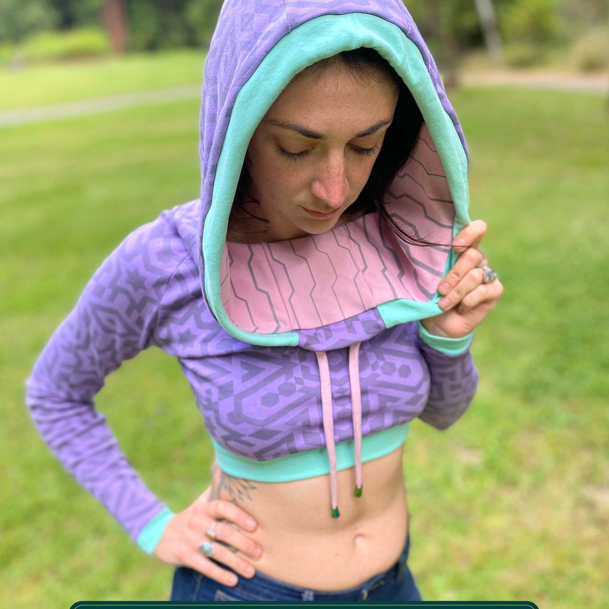 A woman wearing an organic cotton lavender crop hoodie with geometric print and pink accents, worn with cowl hood up, front view.