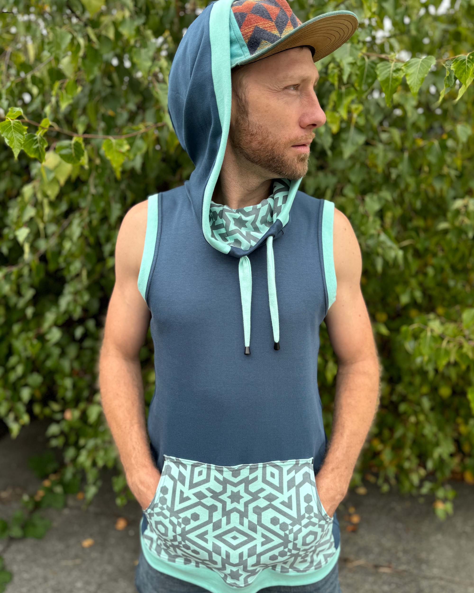 Organic cotton sleeveless hoodie, deep blue with mint geo print accents, front view on male model, hood up.