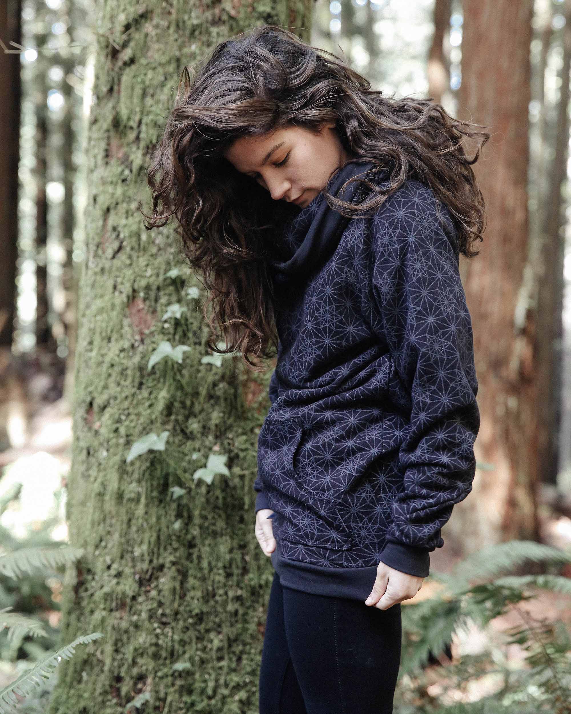 Organic cotton fleece unisex hoodie, black fabric with geometric print, left side view on female model in nature.