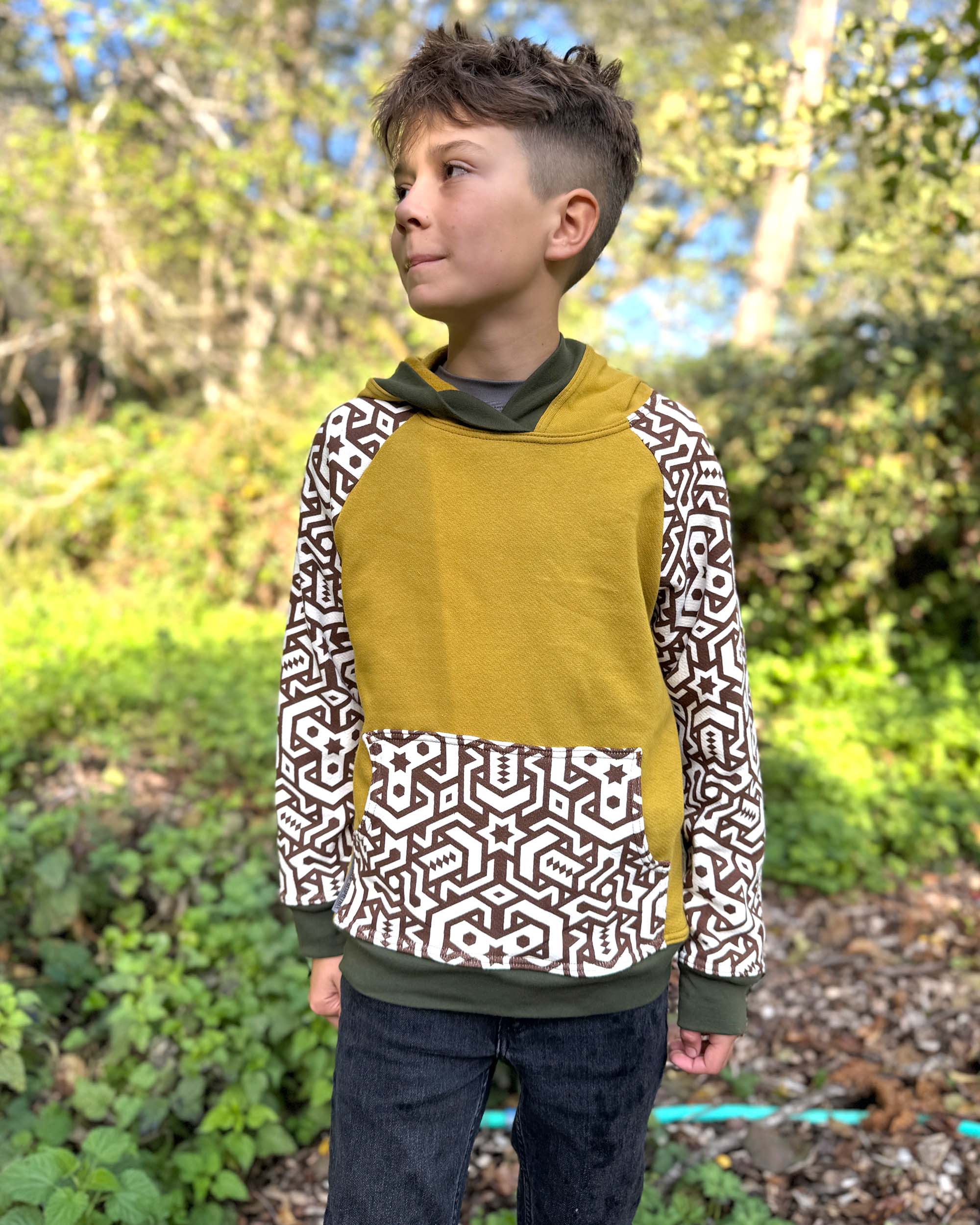 Organic cotton kids hoodie, yellow with geometric printed sleeves, front view, worn by a boy in nature.