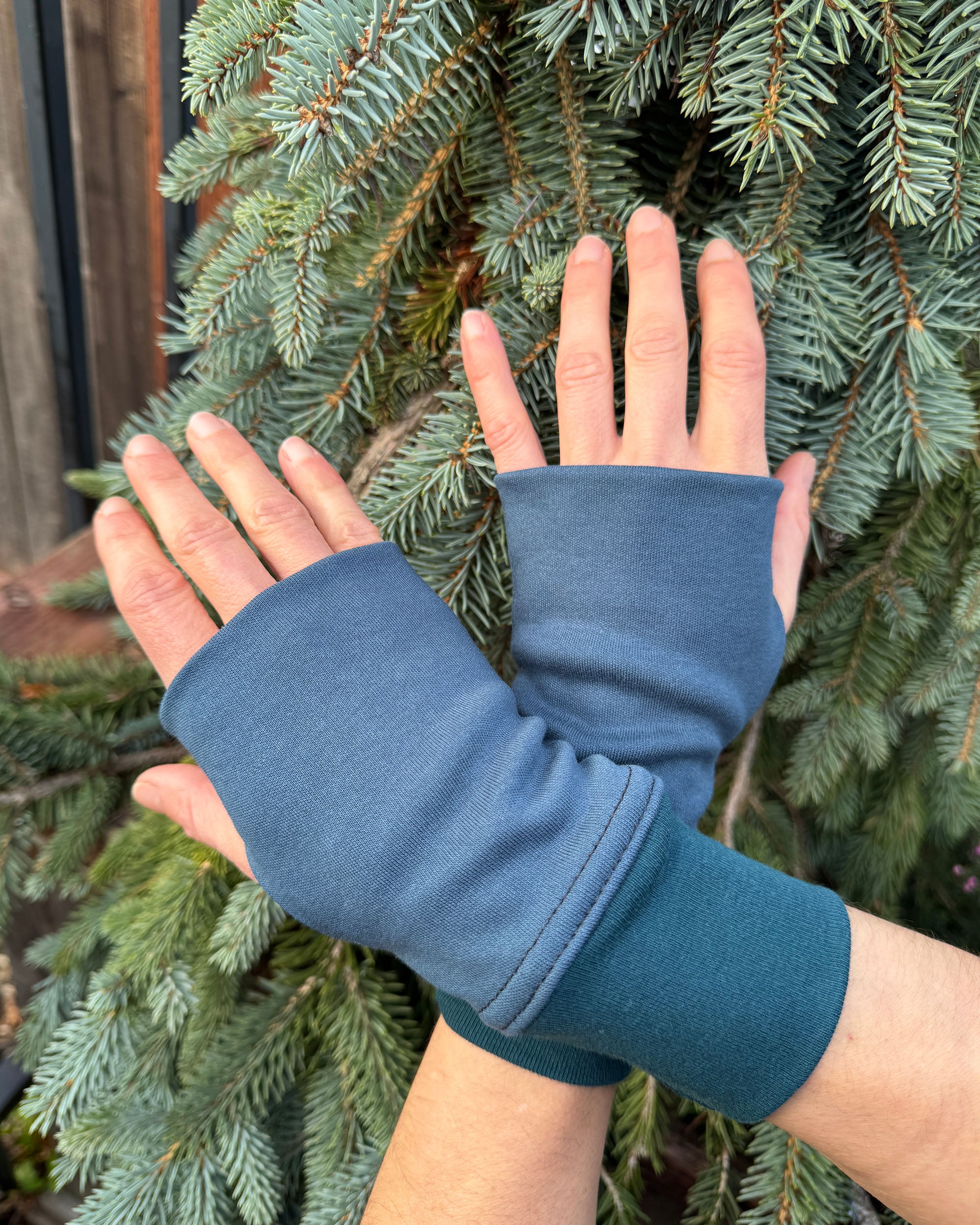 Dark blue organic cotton hand warmers worn by female hands in front of spruce tree.
