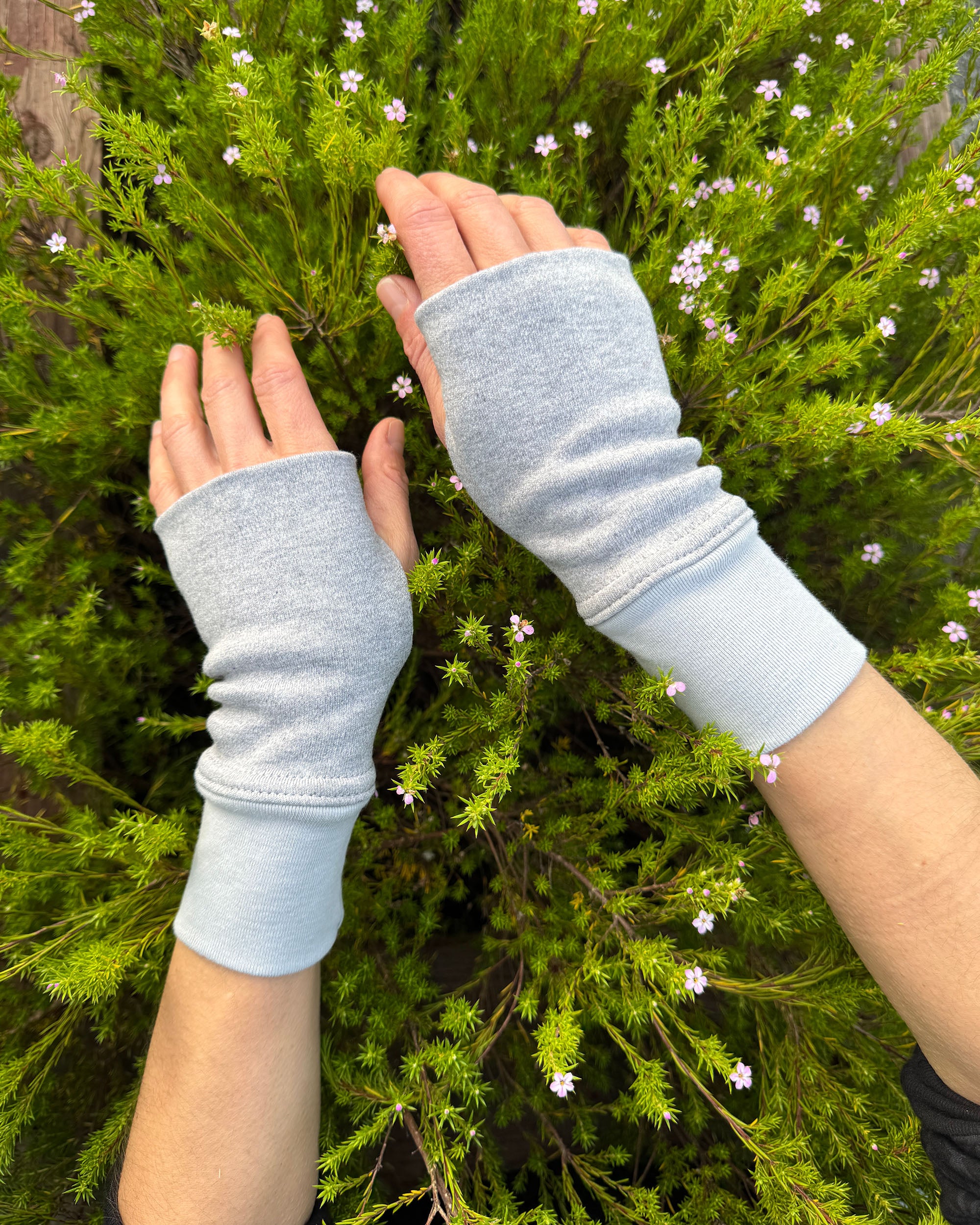 Two hands wearing solid grey organic cotton wrist warmers with a flowery background.