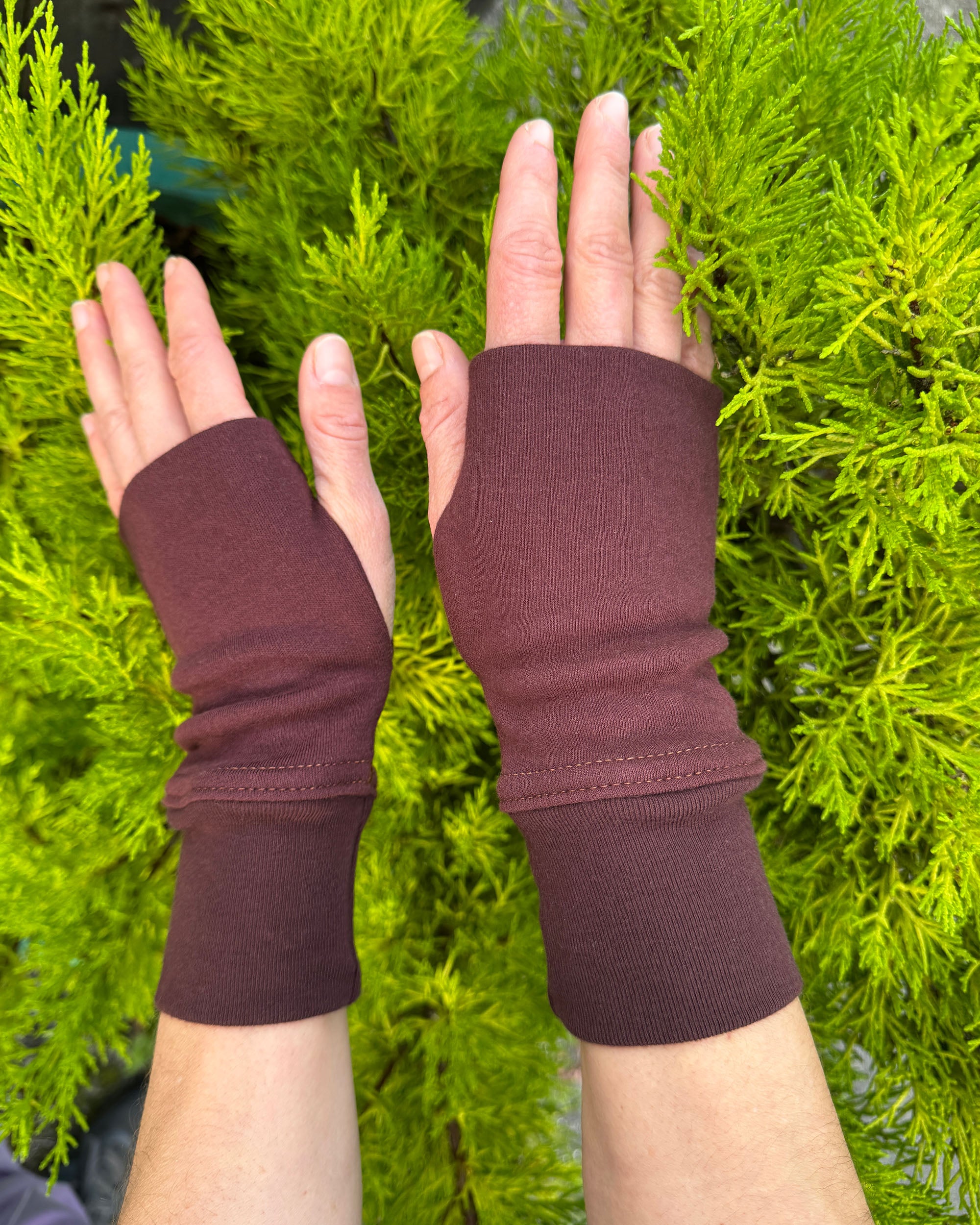 Two hands wearing organic cotton hand warmers, reddish brown colored fabric with natural background.