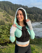 Fitted crop hoodie with cowl hood, mint fabric with black accent, front side view on model on beach.
