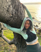 Fitted crop hoodie with cowl hood, mint fabric with black accent, front side view on model with tree.