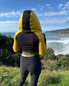 Fitted crop hoodie with cowl hood, yellow fabric with black accent, back side view on model.