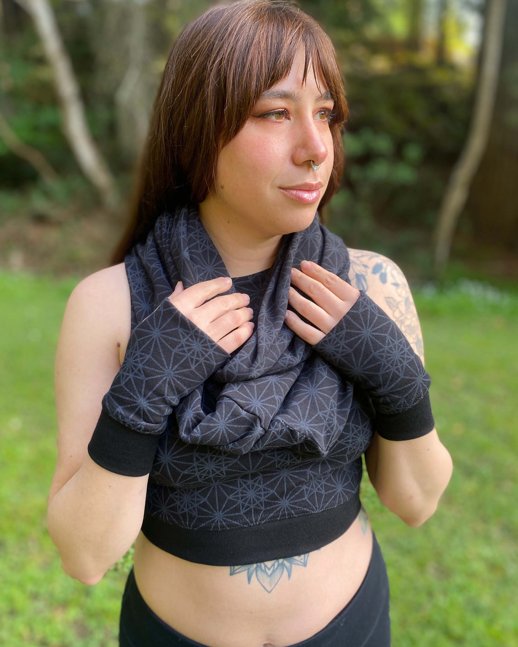 Female model wearing matching set of crop top, infinity scarf and fingerless gloves, black fabric with geometric print, front view in nature.