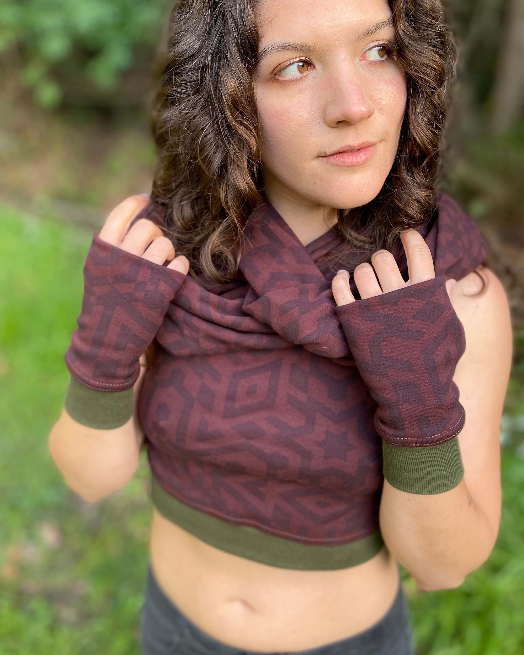 Female model wearing matching set of crop top, infinity scarf and fingerless gloves, black organic cotton fabric with geometric print, front view in nature.
