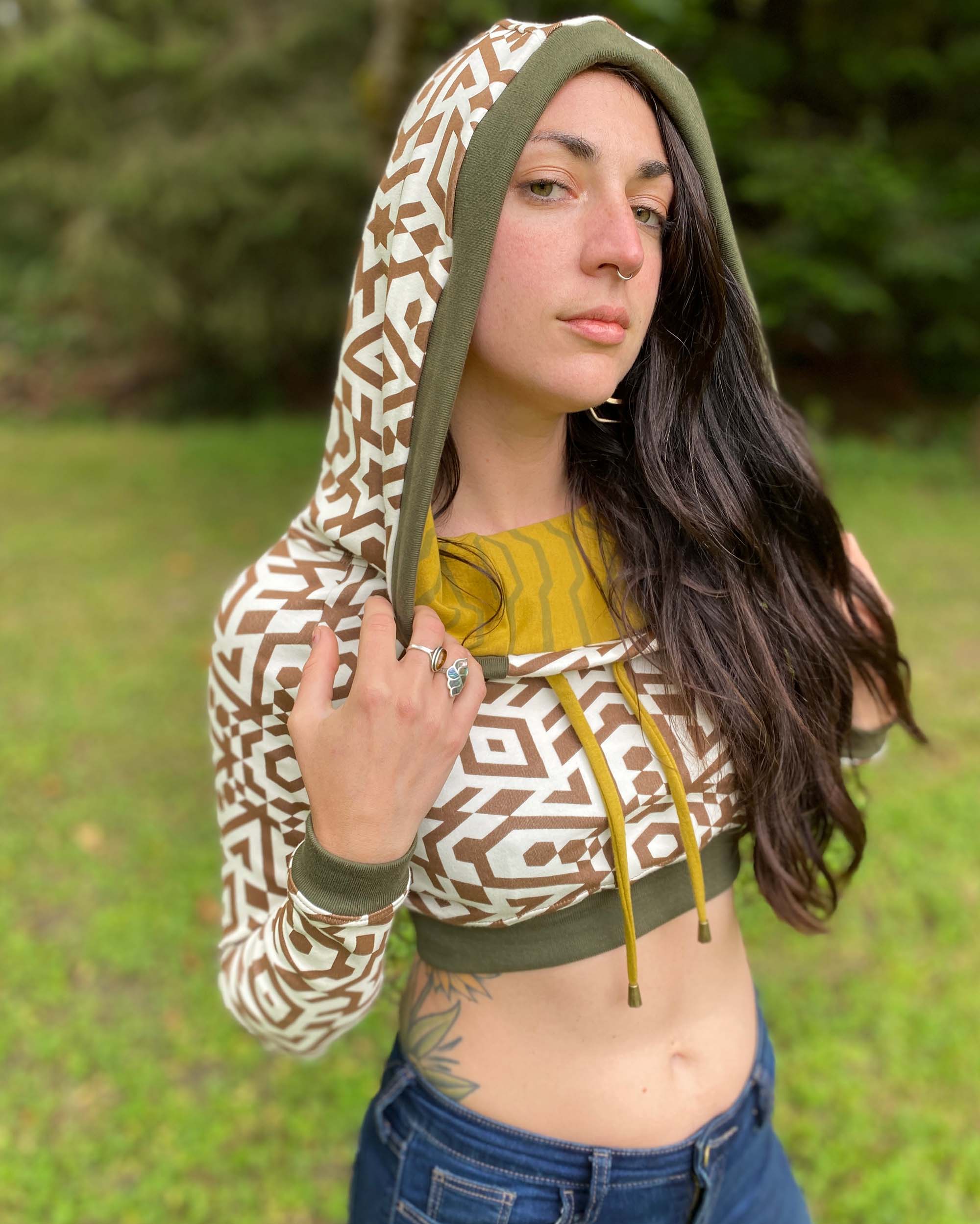 A woman wearing an organic cotton undyed crop hoodie with brown geometric print, worn with hood up, side view.