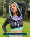 A woman wearing an organic cotton black crop hoodie with geometric print and purple accents, cowl hood and long sleeves, front view with hood up.