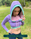 A woman wearing an organic cotton lavender crop hoodie with geometric print and pink accents, worn with cowl hood up, front view.