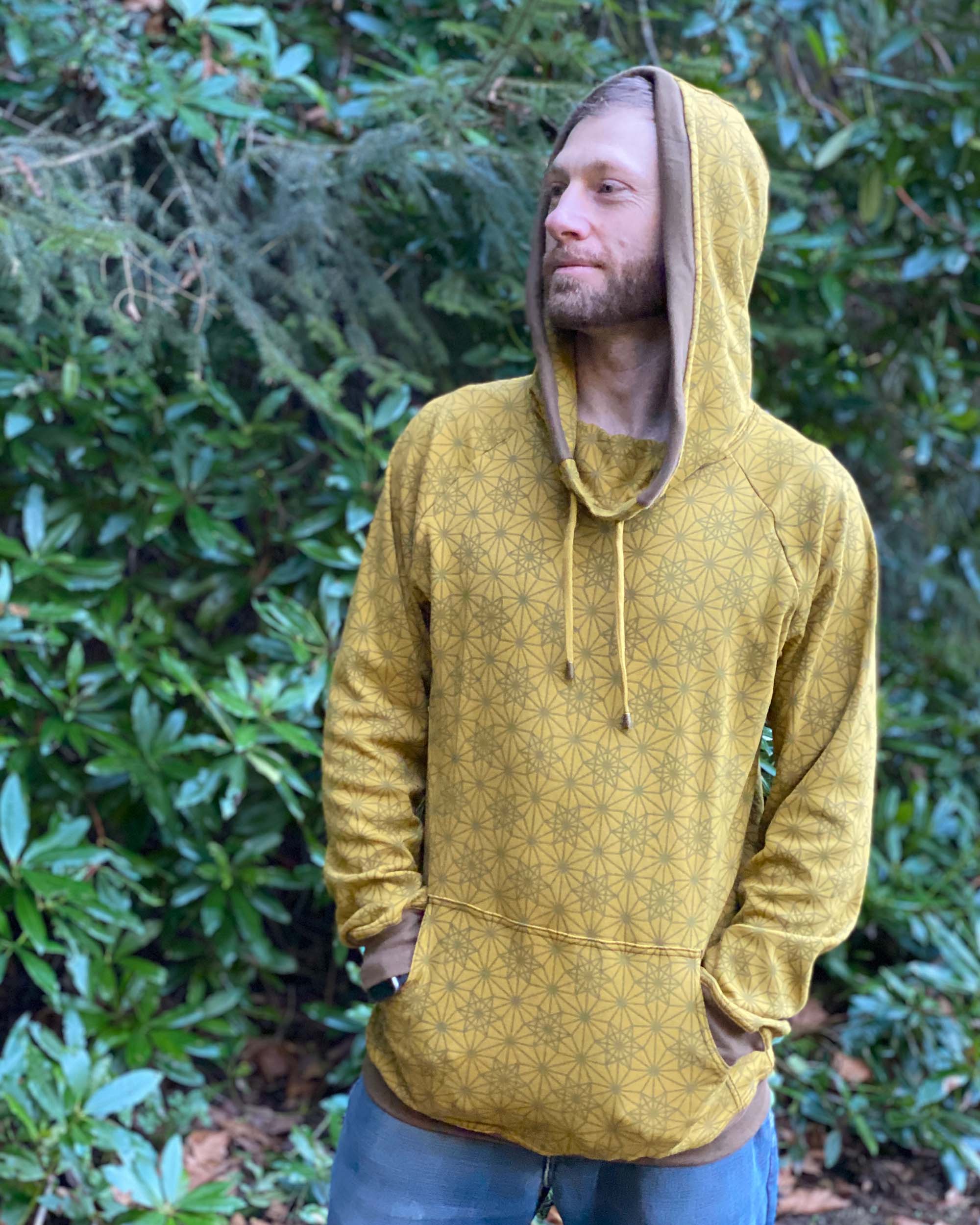 Organic cotton extra long unisex hoodie, yellow fabric with geometric print, left side view of male model in nature.
