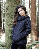Organic cotton fleece unisex hoodie, black fabric with geometric print, front left view on female model in trees.
