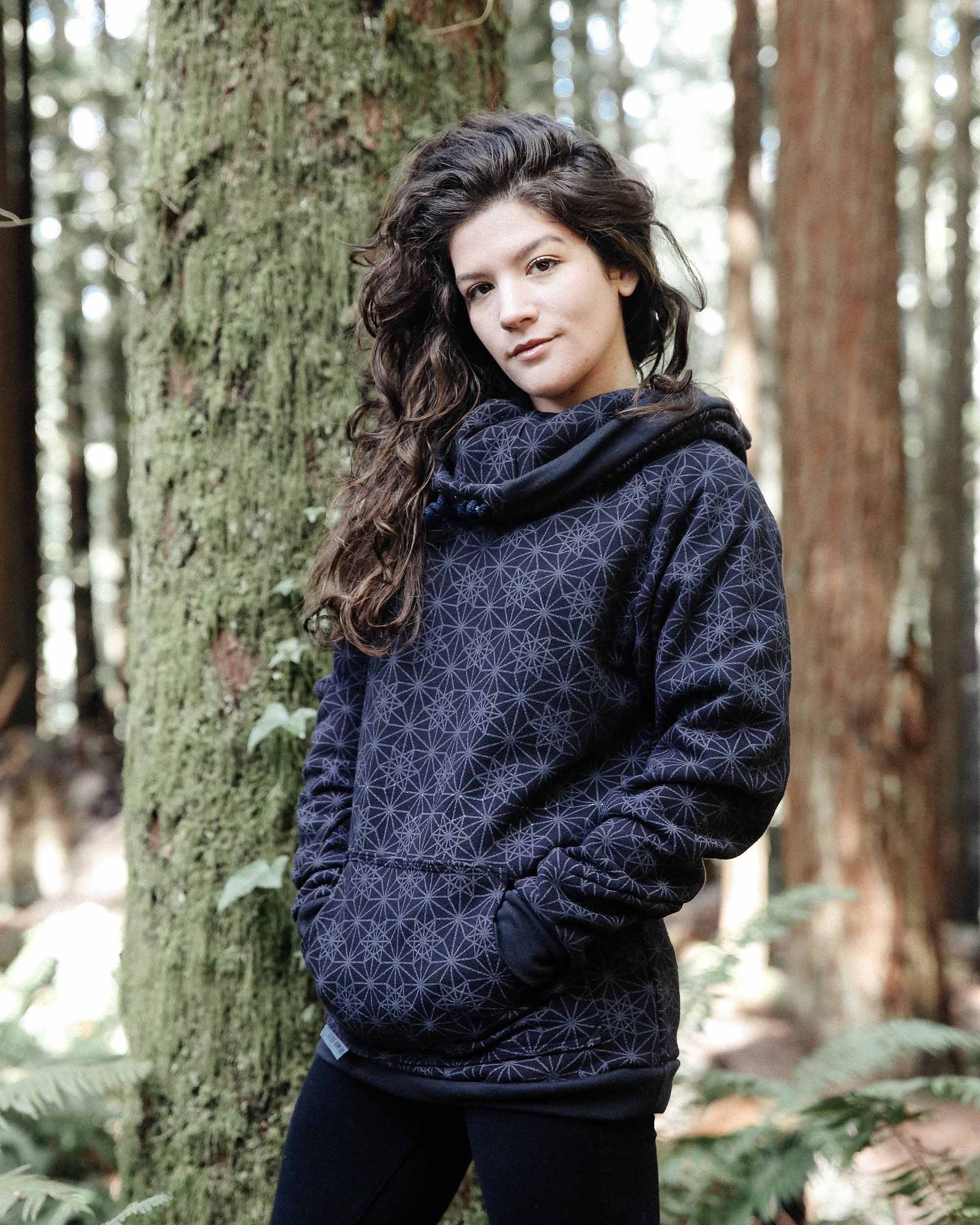 Organic cotton fleece unisex hoodie, black fabric with geometric print, front left view on female model in trees.